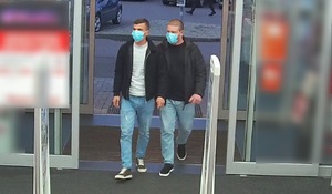 zdjęcie kolorowe: dwóch mężczyzn wchodzących do marketu ze sprzętem elektronicznym. Mężczyzna z lewej ubrany w ciemnozielone buty sportowe z biała podeszwą, niebieskie jeansy, jasnoszara bluzę i czarna kurtkę rozpinana na zamek z założoną niebieska maseczka ochronną na twarzy. Mężczyzna z prawej ubrany w czarne buty sportowe z biała podeszwą, niebieskie jeansy, czarny sweter i czarna kurtkę rozpinaną na zamek z niebieska maseczka ochronna na twarzy