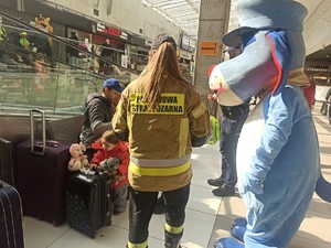 zdjęcie kolorowe: strażaczka z maskotka śląskiej Policji na holu dworca rozmawiająca z rodzicami małych dzieci
