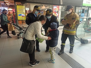 zdjęcie kolorowe: strażniczki miejskie i strażaczka rozdająca elementy odblaskowe rodzicom z dziećmi