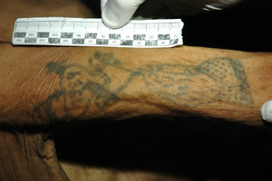 zdjęcie kolorowe: ręka zmarłego mężczyzny z niewyraźnym tatuażem i linijka/wykresówka trzymana przez policjanta