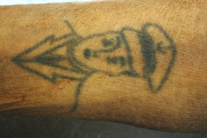 zdjęcie kolorowe: ręka zmarłego mężczyzny i umieszczony na nim tatuaż przedstawiający głowę mężczyzny w czapce