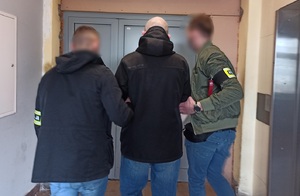 zdjęcie kolorowe: dwóch policjantów wydziału kryminalnego prowadzący mężczyznę poszukiwanego listem gończym