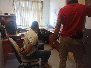 zdjęcie kolorowe: w policyjnym pokoju na krześle obrotowym siedzi zatrzymany mężczyzna z rękami do tyłu, na których założone są kajdanki. Przy zatrzymanym mężczyźnie stoi policjant wydziału kryminalnego