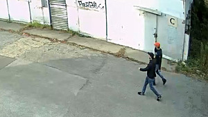 zdjęcie kolorowe: na tle zabudowań gospodarczych dwóch mężczyzn ubranych w ciemne kurtki i jasne spodnie jeansowe. Jeden z nich z czerwoną czapka z daszkiem na głowie