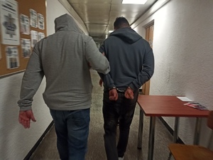 zdjęcie kolorowe:  mężczyzna w ciemnej bluzie dresowej z założonymi na ręce trzymane z tyłu kajdankami prowadzony przez policjanta kryminalnego po korytarzu w katowickiej komendzie,