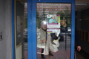zdjęcie kolorowe: katowicki policjant wspólnie z seniorką zawieszają plakat na drzwiach wejściowych do bloku ostrzegający seniorów przed oszustami
