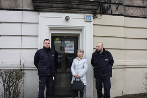 zdjęcie kolorowe: dwóch policjantów z seniorka przed wejściem do budynku w którym rozwieszono plakaty ostrzegające przed oszustami