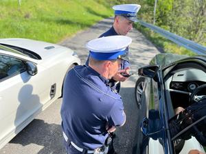 zdjęcie kolorowe: dwóch policjantów ruchu drogowego stojących pomiędzy radiowozem a samochodem osobowym rozmawiający z kierującym pojazdem podczas kontroli drogowej