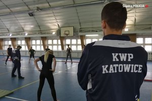 zdjęcie kolorowe: policjant w granatowym dresie z napisem na plecach o treści KWP Katowice, obserwujący osoby ćwiczące na hali sportowej