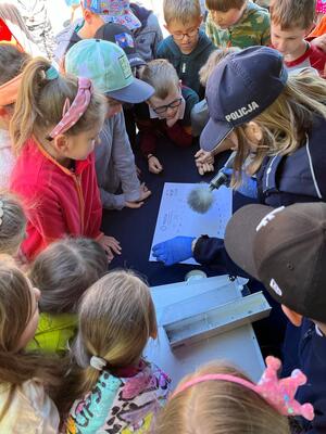 zdjęcie kolorowe: policjantka oraz dzieci zgromadzone wokół stołu, na którym funkcjonariuszka pokazuje dzieciom odciski palców na papierze