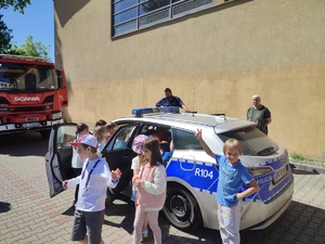 zdjęcie kolorowe: policjant nadzorujący zwiedzanie policyjnego radiowozu przez dzieci zgromadzone na festynie, w tle wóz strażacki
