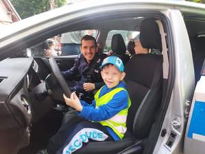 Zdjęcie kolorowe: policjant siedzący w radiowozie z chłopcem, który trzyma kierownicę