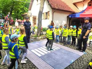 Zdjęcie kolorowe: policjanci z dziećmi ubranymi w odblaskowe kamizelki, które ćwiczą bezpieczne przechodzenie przez przejście dla pieszych