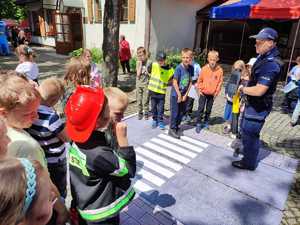 Zdjęcie kolorowe: policjant i  dzieci podczas nauki przechodzenia przez przejście dla pieszych