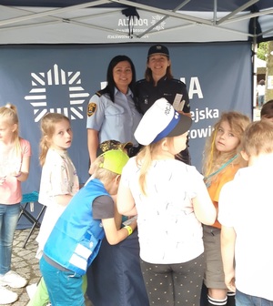 Zdjęcie kolorowe: policjantka i strażniczka miejska na stanowisku profilaktycznym  rozmawiające z dziećmi o bezpieczeństwie w ruchu drogowym