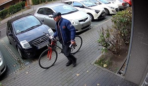 zdjęcie kolorowe: chodnikiem porusza się mężczyzna, który prowadzi skradziony czerwony rower. Mężczyzna ubrany jest w ciemne buty sportowe, ciemne spodnie, ciemna bluzę sportowa i granatowa kurtkę puchową i ciemno szarą czapkę z daszkiem.