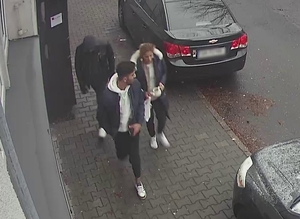 zdjęcie kolorowe: kobieta ubrana w długi puchowy granatowy płaszcz i mężczyzna ubrany w ciemne spodnie, granatowa kurtkę i jasną bluzę dresową z kapturem założonym na głowę oraz mężczyzna ubrany w czarne spodnie, czarna kurtkę i ciemną bluzę dresową z kapturem złożonym na głowę wychodzący z klatki budynku