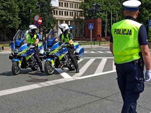 zdjęcie kolorowe: dwóch policjantów katowickiej drogówki na motorach służbowych i policjant kierujący ruchem na skrzyzowaniu