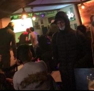 zdjęcie kolorowe: mężczyzna ubrany w czarna kurtkę z kapturem z maseczka założoną na twarzy stojący w lokalu gastronomicznym