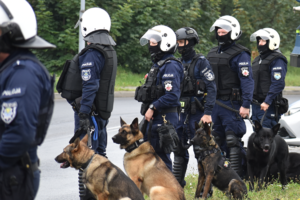 Zdjęcie kolorowe przedstawia policjantów przewodników psów służbowych na tle armatki wodnej.