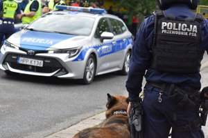 Zdjęcie kolorowe przedstawia policjanta przewodnika psa służbowego na tle radiowozu policyjnego.