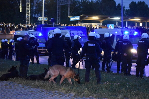 Zdjęcie kolorowe przedstawia policjantów zabezpieczających teren przyległy do stadionu piłkarskiego.