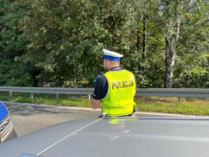 Zdjęcie kolorowe przedstawia policjanta z drogówki mierzącego prędkość pojazdów.