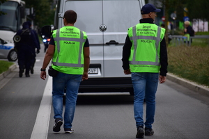zdjęcie kolorowe: dwóch policjantów zespołu antykonfliktowego idących za białym samochodem