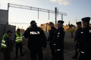 Zdjęcie kolorowe przedstawia umundurowanych policjantów w trakcie czynności służbowych na obszarze kolejowym.