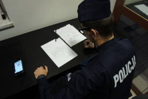 Zdjęcie kolorowe przedstawia umundurowanego policjanta podczas sporządzania dokumentacji służbowej w jednostce Policji.