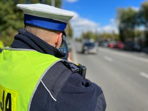 Zdjęcie kolorowe przedstawia umundurowanego policjanta dokonującego pomiaru prędkości jadącego pojazdu.