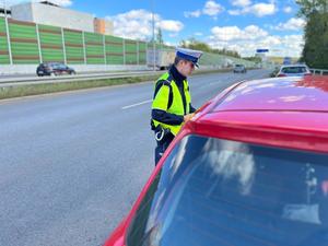 Zdjęcie kolorowe przedstawia umundurowanego policjanta drogówki, który wykonuje kontrolę pojazdu oraz kierującego.
