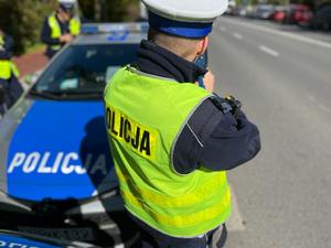 Zdjęcie kolorowe przedstawia umundurowanego policjanta Wydziału Ruchu Drogowego dokonującego pomiaru prędkości jadącego pojazdu.