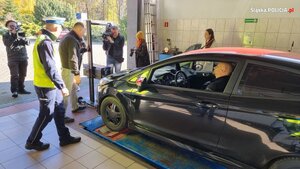 zdjęcie kolorowe: policjant na stacji diagnostycznej przyglądający się badaniu świateł w samochodzie osobowym w obecności kamer telewizyjnych