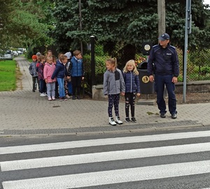 zdjęcie kolorowe: umundurowany policjant z przedszkolakami stojący przy przejściu dla pieszych, który sprawdza czy bezpiecznie można przejść przez jezdnię