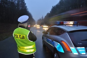 zdjęcie kolorowe: policjantka wydziału ruchu drogowego stojąca przy radiowozie, która nadzoruje przejazd samochodów