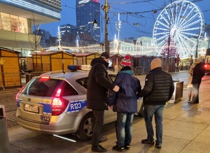 zdjęcie kolorowe: dwóch policjantów stojących z zatrzymanym przed radiowozem na katowickim rynku