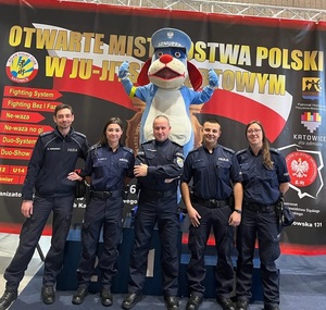 zdjęcie kolorowe: 5 katowickich policjantów i maskotka śląskiej Policji Sznupek na tle baneru Turnieju Młodych Mistrzów w Ju-Jitsu Sportowym