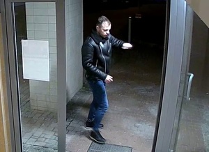 zdjęcie kolorowe: mężczyzna ubrany w granatowe spodnie i czarna kurtkę stojący przed otwartymi drzwiami klatki schodowej