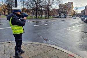 Zdjęcie kolorowe przedstawia policjanta z drogówki mierzącego prędkość poruszającego się pojazdu.