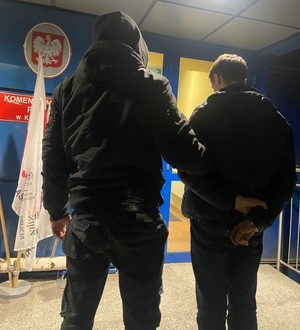 zdjęcie kolorowe: policjant katowickiej komendy przed budynkiem Policji trzymający za ramie mężczyznę zatrzymanego za kradzież katalizatorów