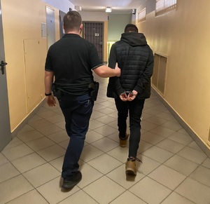 zdjęcie kolorowy: korytarz w policyjnym areszcie. młody Mężczyzna ubrany w czarne spodnie i czarną kurtkę z rękami na które założono kajdanki prowadzony pod ramie przez policjanta