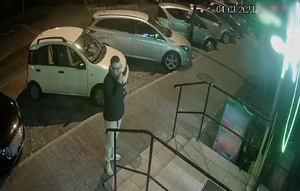 zdjęcie kolorowe: na chodniku przed sklepem stoi mężczyzna ubrany w jasne buty sportowe jasne spodnie dresowe i czarną kurtkę. Na twarzy założone okulary korekcyjne