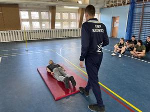 zdjęcie kolorowe: uczeń szkoły średniej wykonujący ćwiczenie na macie w policyjnej sali gimnastycznej pod okiem instruktora