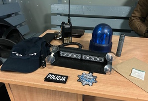 zdjęcie kolorowe: na biurku ułożono czapkę z daszkiem z napisem „Policja”, lampę błyskowa koloru niebieskiego tzw. „kogut”, przenośną radiostację, naszywkę z rzepem i napisem „Policja”, a także haftowana na wzór odznaki policyjnej naszywkę z napisem „Policja” oraz numerem identyfikacyjnym, a także woreczek foliowy z zawartością białej sypkiej substancji