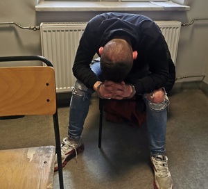 zdjęcie kolorowe: młody mężczyzna ubrany w niebieskie spodnie i granatowy sweter siedzący na krześle, gdzie na ręce trzymane z przodu założono mu kajdanki