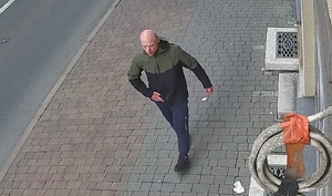 zdjęcie kolorowe: łysy mężczyzna ubrany w czarne buty sportowe, granatowe spodnie dresowe i czarno-zieloną kurtkę idzie chodnikiem