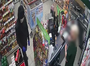 zdjęcie kolorowe: ubrany na czarno mężczyzna, w bluzie z kapturem  założonym na głowę, z zamaskowana twarzą stoi prze sklepowej ladzie i rozmawia z ekspedientką