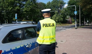 zdjęcie kolorowe: policjant ruchu drogowego w rejonie przejścia dla pieszych stojący przy radiowozie