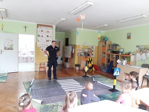 zdjęcie kolorowe: sala w przedszkolu. Policjant rozmawiający z dziećmi o bezpiecznym przechodzeniu przez przejście dla pieszych